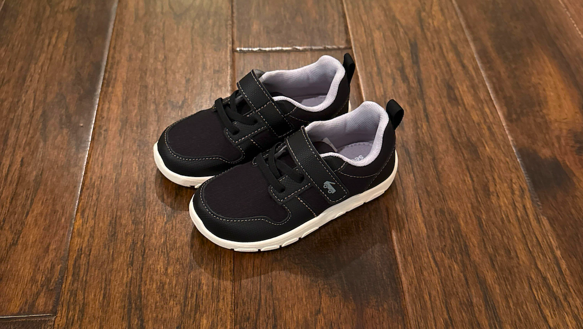 Black toddler sneakers with white soles on wooden floor, featuring Velcro strap and soft inner lining for comfort.
